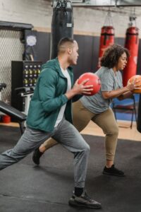 Side view of fit young ethnic male couch instructing focused obese African American lady doing lunges with medicine ball in gym